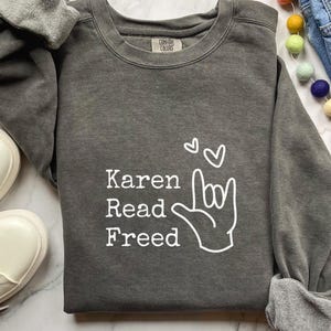 Puede incluir: Sudadera gris oscuro con el texto "Karen Read Freed" y un gráfico de gesto de la mano. La sudadera está hecha de un material suave y tiene un ajuste relajado. Zapatillas blancas y vaqueros azules son visibles en el fondo.