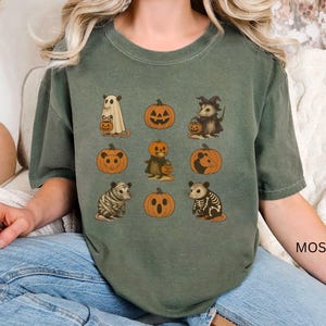 K&ouml;nnte beinhalten: Moosgr&uuml;nes T-Shirt mit Halloween-Illustrationen. Das Design umfasst einen Geist, K&uuml;rbisse und verschiedene Tiere wie ein Opossum und einen Tiger, die alle K&uuml;rbislaternen halten. Das Wort "MOSS" ist auf dem &Auml;rmel aufgedruckt.