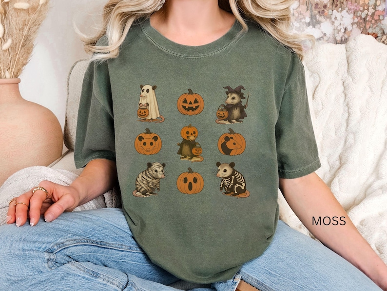K&ouml;nnte beinhalten: Moosgr&uuml;nes T-Shirt mit Halloween-Illustrationen. Das Design umfasst einen Geist, K&uuml;rbisse und verschiedene Tiere wie ein Opossum und einen Tiger, die alle K&uuml;rbislaternen halten. Das Wort "MOSS" ist auf dem &Auml;rmel aufgedruckt.