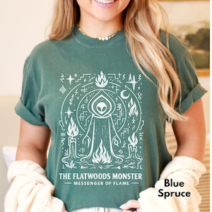 May include: Blue Spruce t-shirt with a white graphic of the Flatwoods Monster, surrounded by flames, candles, and celestial symbols. The text "THE FLATWOODS MONSTER - MESSENGER OF FLAME" is printed below the graphic. The shirt is a comfortable, casual style.
