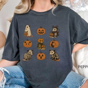 K&ouml;nnte beinhalten: Dunkelgraues T-Shirt mit Halloween-Illustrationen. Das Design zeigt einen Geist, K&uuml;rbisse und verkleidete Opossums. Das Wort "PEPPER" ist auf der unteren rechten Seite des Shirts aufgedruckt.
