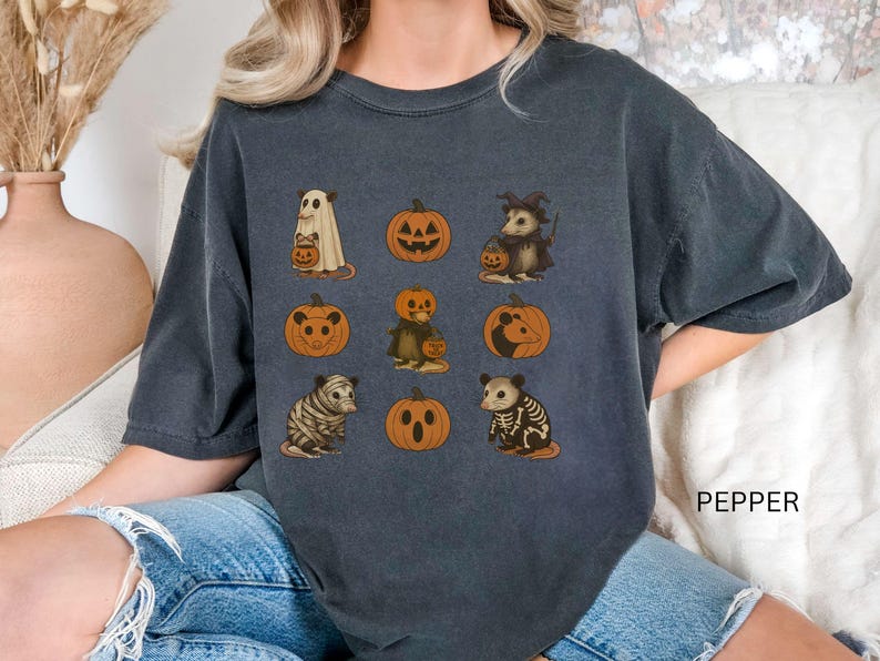 K&ouml;nnte beinhalten: Dunkelgraues T-Shirt mit Halloween-Illustrationen. Das Design zeigt einen Geist, K&uuml;rbisse und verkleidete Opossums. Das Wort "PEPPER" ist auf der unteren rechten Seite des Shirts aufgedruckt.