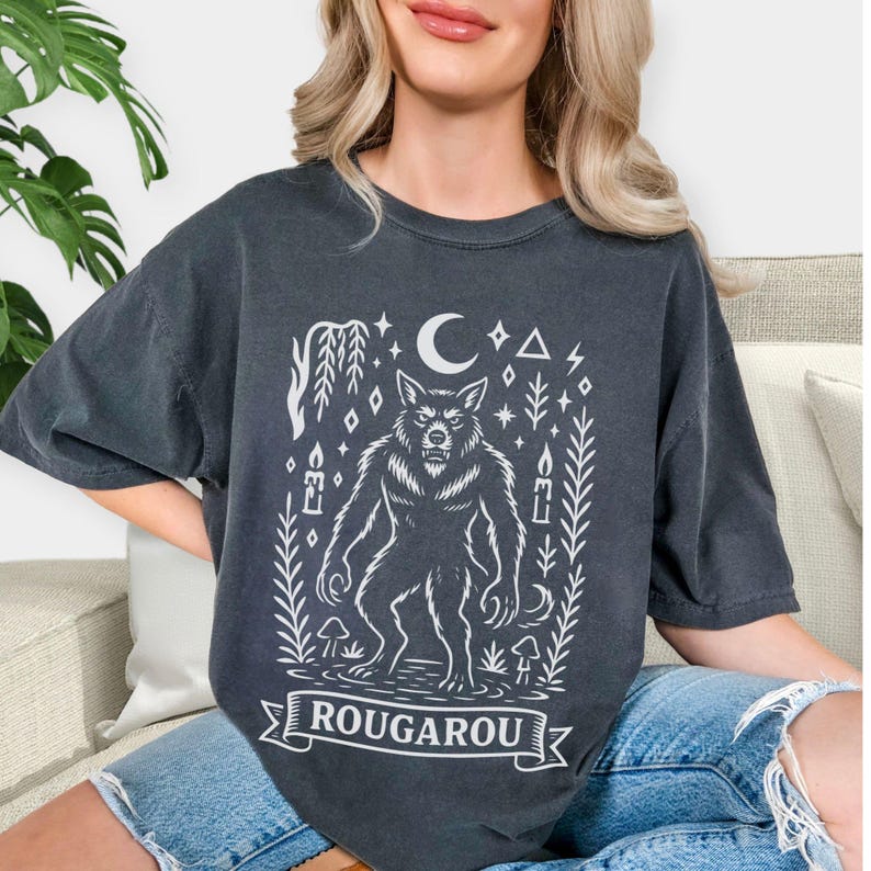 May include: Dark gray t-shirt featuring a white illustration of a werewolf, surrounded by a crescent moon, stars, and botanical elements. The word "ROUGAROU" is displayed on a banner at the bottom. The shirt has a relaxed fit.