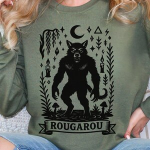 May include: Olive green sweatshirt featuring a black Rougarou graphic. The design includes a werewolf, a crescent moon, stars, candles, and the word "ROUGAROU" on a banner. The sweatshirt has a crew neck and long sleeves.