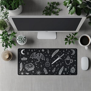May include: Black rectangular desk mat with a white fantasy-themed design. The design includes a dragon, sword, feathers, stars, a crescent moon, a crown, a book, and a steaming mug. A computer monitor, coffee cup, and mouse are also visible.