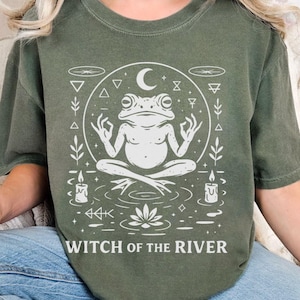 May include: Olive green t-shirt featuring a white graphic of a meditating frog within a circle, with a crescent moon, candles, and the text "WITCH OF THE RIVER". The design has a mystical, spiritual theme.