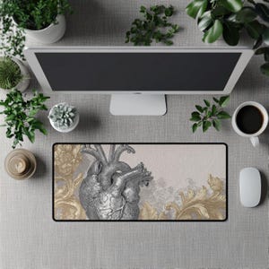Puede incluir: Alfombrilla de ratón rectangular con una detallada ilustración de un corazón anatómico plateado sobre un fondo beige con detalles florales dorados. La alfombrilla tiene un borde negro. También se ven un monitor de ordenador, una taza de café y un ratón.