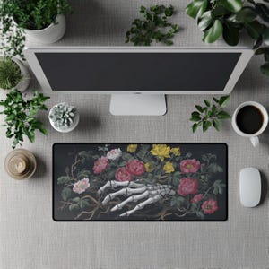 Puede incluir: Un ratón rectangular negro con una mano esquelética que se extiende hacia un ramo de flores rosas, rojas y amarillas. El diseño floral está sobre un fondo oscuro. El ratón está rodeado por un ordenador, plantas y una taza de café.