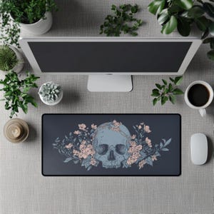 Puede incluir: Alfombrilla de ratón rectangular con un fondo gris oscuro que presenta un diseño de calavera azul claro adornado con flores rosas y hojas verdes. También se ven un monitor de ordenador, una taza de café y un ratón.