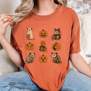 K&ouml;nnte beinhalten: Gebrannt orangefarbenes T-Shirt mit Halloween-Illustrationen. Das Design zeigt Tiere und K&uuml;rbisse. Die Tiere sind in Halloween-Kost&uuml;men gekleidet, darunter ein Geist, eine Hexe und ein Skelett. Das Wort "YAM" ist auf der rechten Seite des Shirts aufgedruckt.