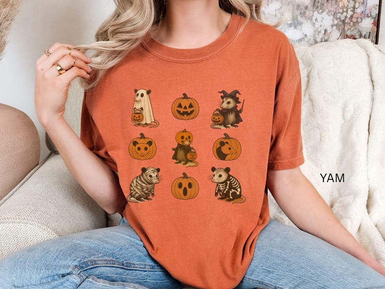 K&ouml;nnte beinhalten: Gebrannt orangefarbenes T-Shirt mit Halloween-Illustrationen. Das Design zeigt Tiere und K&uuml;rbisse. Die Tiere sind in Halloween-Kost&uuml;men gekleidet, darunter ein Geist, eine Hexe und ein Skelett. Das Wort "YAM" ist auf der rechten Seite des Shirts aufgedruckt.