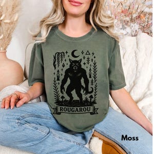 May include: Moss green t-shirt featuring a black Rougarou graphic. The design includes a werewolf, a crescent moon, candles, and the word "Rougarou" on a banner. The shirt is a crew neck and has short sleeves.