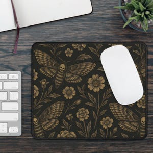 May include: Black rectangular mousepad with a detailed gold illustration of a moth with a skull design, set against a floral background. A white computer mouse sits on the pad, with a keyboard and notebook visible.