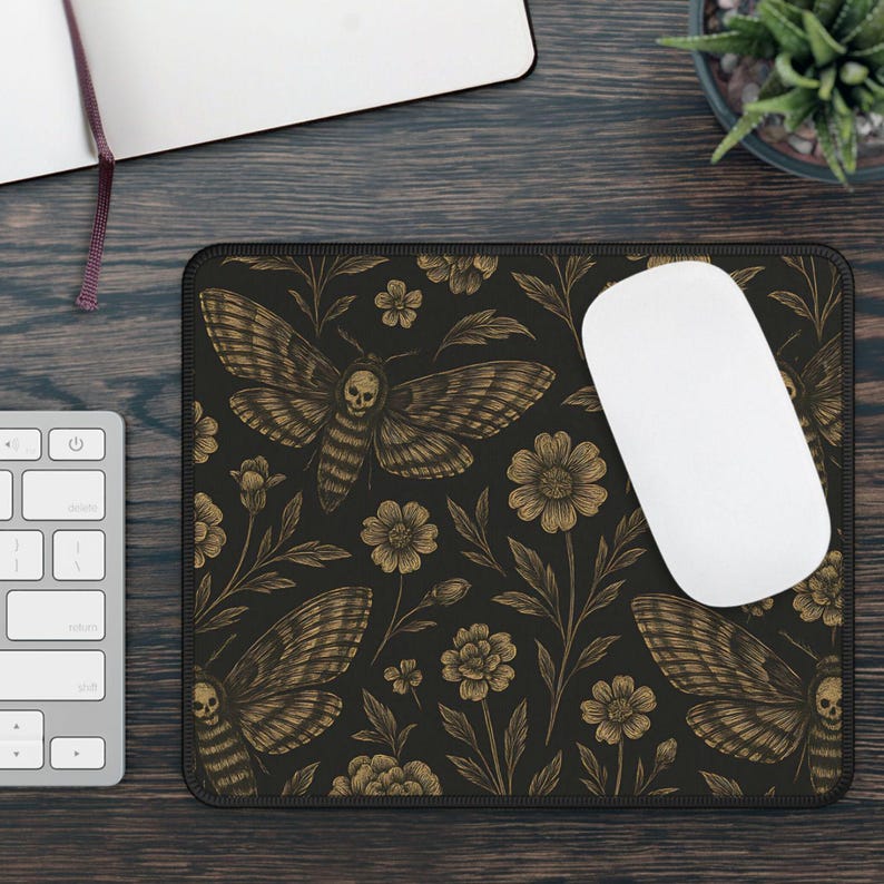 May include: Black rectangular mousepad with a detailed gold illustration of a moth with a skull design, set against a floral background. A white computer mouse sits on the pad, with a keyboard and notebook visible.