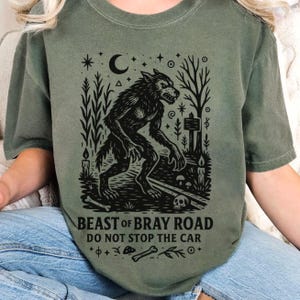 Op de afbeelding: Olijfgroen t-shirt met een zwarte afbeelding van een weerwolf onder een maan, omringd door bomen, kaarsen en een schedel. De tekst "BEAST OF BRAY ROAD DO NOT STOP THE CAR" staat eronder.