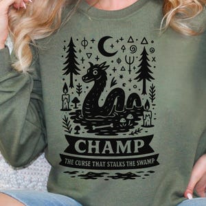 May include: Olive green sweatshirt with a black graphic of a lake monster, trees, candles, and a crescent moon. The word "CHAMP" is printed on a banner, with the text "THE CURSE THAT STALKS THE SWAMP" below.