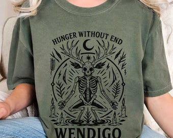 May include: Olive green t-shirt featuring a black graphic of a skeleton with antlers, surrounded by a circular design with the text "HUNGER WITHOUT END" above and "WENDIGO" below. The design includes a crescent moon, star, and candles.