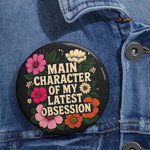 Puede incluir: Un botón negro con un diseño floral y el texto "Main Character of My Latest Obsession".