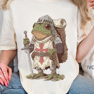 Può includere: T-shirt color avorio con un'illustrazione dettagliata di un cavaliere rana. La rana è vestita con un'armatura, tiene una spada e indossa un elmo. Uno zaino è legato alla sua schiena. Il design include una croce rossa sulla tunica della rana.
