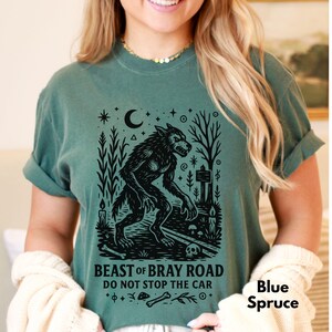 Op de afbeelding: Blauwspar T-shirt met een zwarte weerwolfafbeelding en de tekst "BEAST OF BRAY ROAD DO NOT STOP THE CAR". Het ontwerp bevat een maan, sterren, bomen en een schedel. Het shirt is effen van kleur.