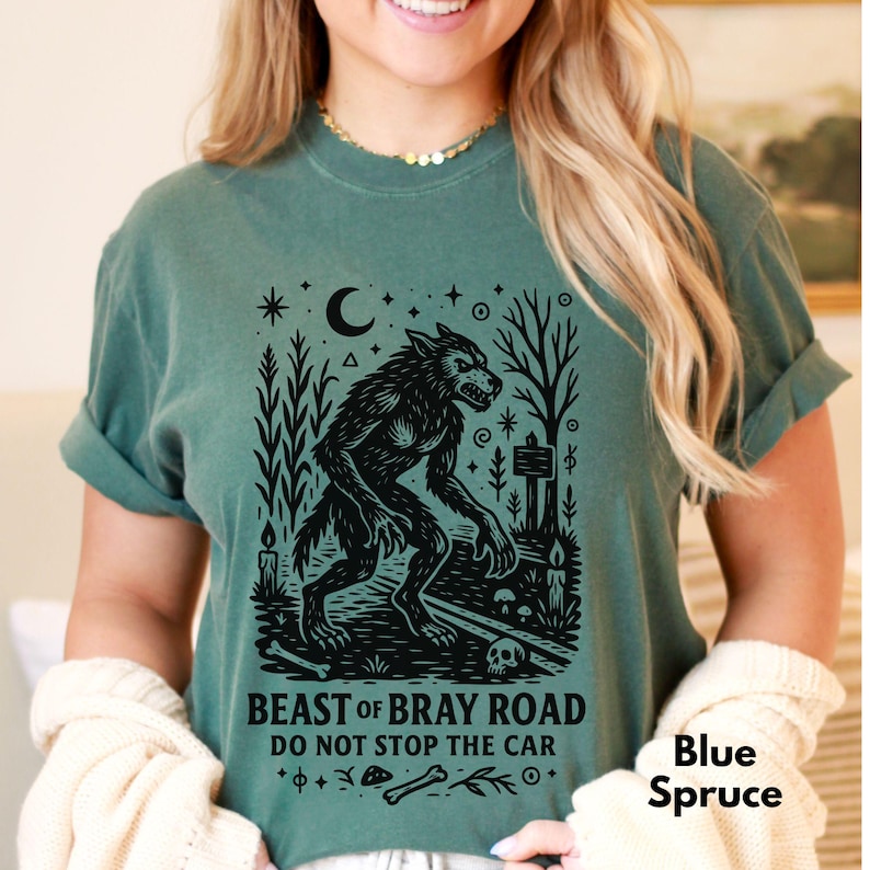 Op de afbeelding: Blauwspar T-shirt met een zwarte weerwolfafbeelding en de tekst "BEAST OF BRAY ROAD DO NOT STOP THE CAR". Het ontwerp bevat een maan, sterren, bomen en een schedel. Het shirt is effen van kleur.