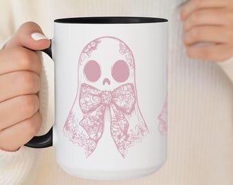 May include: White ceramic mug with a black interior and handle. The mug features a pink illustration of a ghost with a skull face and a large bow. The ghost design is detailed with floral lace patterns.