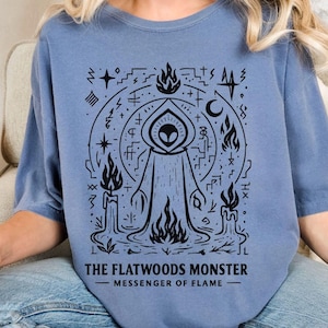 May include: A blue-grey t-shirt featuring a black graphic of the Flatwoods Monster, surrounded by flames, candles, and symbols. The text "THE FLATWOODS MONSTER - MESSENGER OF FLAME" is printed below the graphic.