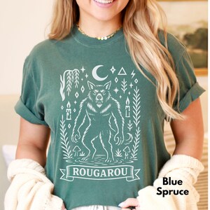 May include: Blue spruce t-shirt with a white graphic of a werewolf, moon, stars, and the word "ROUGAROU" on a banner. The shirt has rolled-up sleeves and a comfortable fit. The text "Blue Spruce" is also visible.