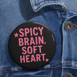 May include: A black button with a pink star and the text "SPICY BRAIN, SOFT HEART" in pink.