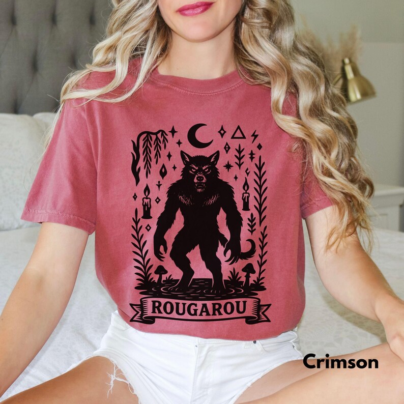 May include: Crimson-colored t-shirt featuring a black Rougarou graphic. The design includes a werewolf, crescent moon, candles, and the word "ROUGAROU" on a banner. The shirt is a short-sleeved, crew neck style.