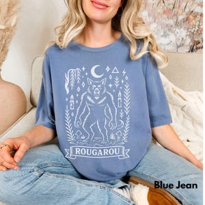 May include: Blue short-sleeved t-shirt with a white graphic of a werewolf, moon, and stars. The word "ROUGAROU" is displayed on a banner. The text "Blue Jean" is at the bottom right.