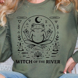 May include: Sage green sweatshirt featuring a black graphic of a meditating frog inside a circle with a crescent moon and other symbols. The text "WITCH OF THE RIVER" is printed below the frog. The sweatshirt has long sleeves.