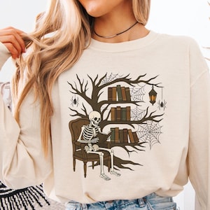 Op de afbeelding: Crèmekleurige sweatshirt met een skelet dat een boek leest in een bruine fauteuil. Achter het skelet staat een boom met boekenplanken, spinnenwebben en een hangende lantaarn. Het ontwerp heeft een Halloween- of gothic-thema.
