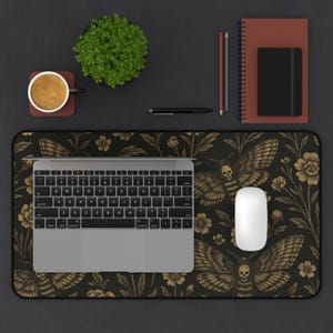 May include: A black desk mat featuring a gold moth and floral pattern, a laptop, and a white mouse. A coffee cup, a small plant, a pen, a pencil, and a notebook are also on the desk. The desk is black.