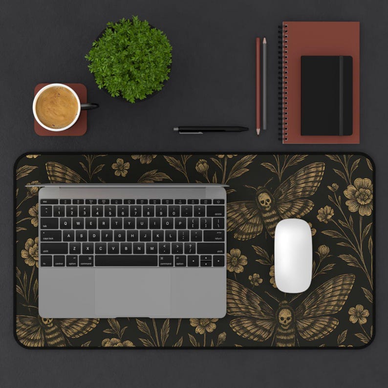 May include: A black desk mat featuring a gold moth and floral pattern, a laptop, and a white mouse. A coffee cup, a small plant, a pen, a pencil, and a notebook are also on the desk. The desk is black.