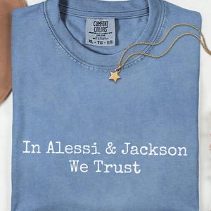 Puede incluir: Una camiseta Comfort Colors azul claro con el texto blanco "In Alessi & Jackson We Trust". Un collar con un colgante de estrella dorada descansa sobre la camiseta. Un sombrero marrón y flores secas están en el fondo.