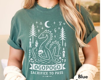 May include: Blue Spruce t-shirt with a white graphic of a lake monster holding a skull, surrounded by trees, candles, and the text "OGOPOGO SACRIFICE TO PASS". The shirt is a solid color.