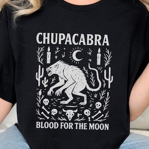 May include: Black t-shirt with a white graphic featuring the word "CHUPACABRA" above an illustration of a creature, a crescent moon, candles, and the text "BLOOD FOR THE MOON".