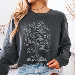 Puede incluir: Una sudadera gris carbón de manga larga con un gráfico de líneas blancas que muestra un árbol con libros en sus ramas, una linterna colgante, un gato durmiendo, una calabaza y setas. El texto "Falling for Fiction" está impreso en color blanco.
