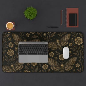 May include: A black desk mat with a gold moth and floral design. A laptop, mouse, and keyboard are on the mat. A small plant, coffee cup, notebook, and pen are also on the desk.