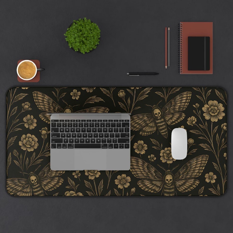 May include: A black desk mat with a gold moth and floral design. A laptop, mouse, and keyboard are on the mat. A small plant, coffee cup, notebook, and pen are also on the desk.