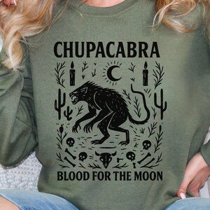 May include: Olive green sweatshirt with a black graphic featuring a Chupacabra, a crescent moon, candles, cacti, and the text "CHUPACABRA BLOOD FOR THE MOON". The design includes skulls, bones, and a bull skull.