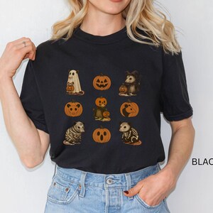 K&ouml;nnte beinhalten: Schwarzes T-Shirt mit Halloween-Illustrationen. Das Design umfasst einen Geist, K&uuml;rbisse und verschiedene Tiere in Kost&uuml;men. Das Wort "BLACK" ist unten rechts auf dem T-Shirt aufgedruckt.