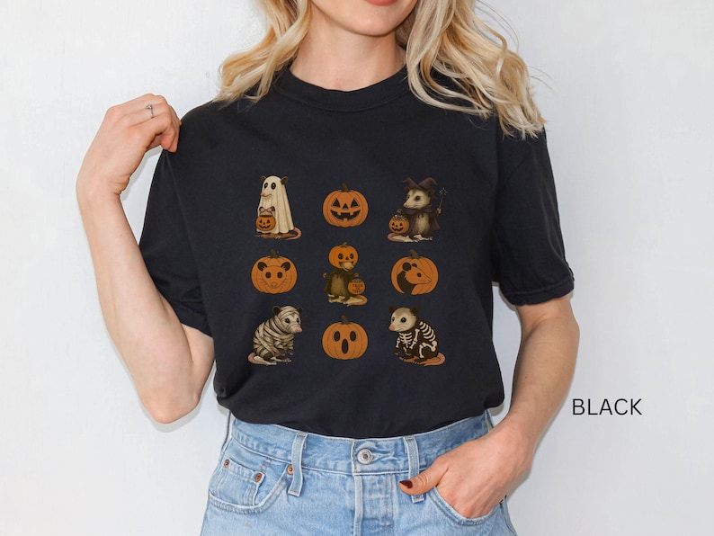 K&ouml;nnte beinhalten: Schwarzes T-Shirt mit Halloween-Illustrationen. Das Design umfasst einen Geist, K&uuml;rbisse und verschiedene Tiere in Kost&uuml;men. Das Wort "BLACK" ist unten rechts auf dem T-Shirt aufgedruckt.