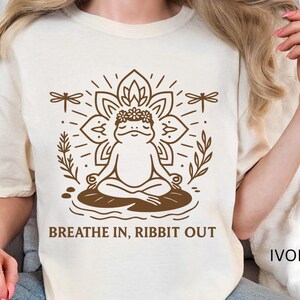 May include: Ivory-colored t-shirt featuring a brown illustration of a meditating frog with a floral crown, surrounded by a lotus flower and dragonflies. The text "BREATHE IN, RIBBIT OUT" is below the frog. The word "IVORY" is in the bottom right corner.