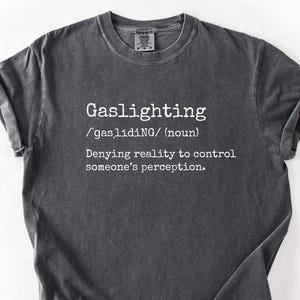 May include: Dark gray t-shirt with the word "Gaslighting" in white, along with its pronunciation and definition. The shirt has a crew neck and short sleeves. The fabric appears to be pre-washed for a vintage look.