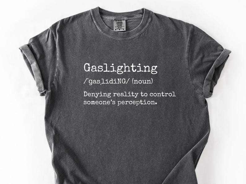 May include: Dark gray t-shirt with the word "Gaslighting" in white, along with its pronunciation and definition. The shirt has a crew neck and short sleeves. The fabric appears to be pre-washed for a vintage look.