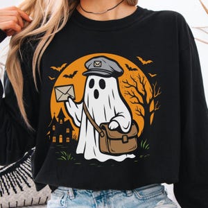 May include: Black sweatshirt with a cartoon ghost postman design. The ghost is holding an envelope and a satchel, set against a Halloween backdrop. The design includes bats, a full moon, and a haunted house, perfect for the spooky season.