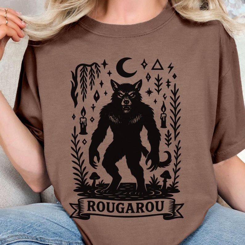 May include: Brown t-shirt featuring a black Rougarou graphic. The design includes a werewolf, a crescent moon, stars, candles, and the word "ROUGAROU" on a banner. The shirt is made of a soft material.
