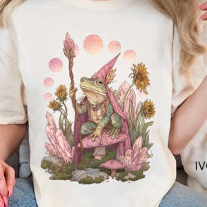 Puede incluir: Camiseta de color marfil con una ilustración fantástica de un mago rana. La rana lleva una capa y un sombrero rosas, sostiene un bastón y está sentada en una seta. El diseño incluye cristales, flores y esferas rosas.